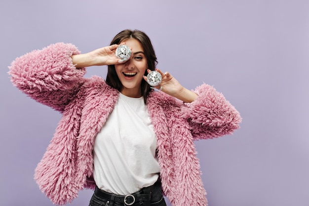 Mujer bastante moderna con peinado oscuro en suéter rosa esponjoso de moda y top blanco sonriendo y posando con bolas de discoteca