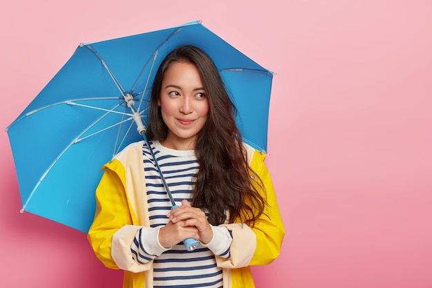 Mujer bastante pensativa con cabello lacio oscuro, sostiene paraguas azul, camina durante el día fresco, se protege de la lluvia