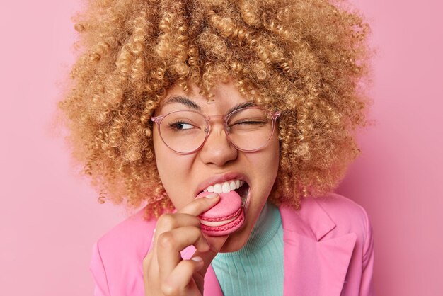 Una mujer bastante rizada muerde hambrienta macarrón dulce guiña los ojos mira hacia algún lado tiene adicción al azúcar usa gafas y ropa formal aislada sobre fondo rosa Deliciosa confitería