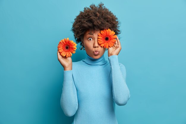 mujer bastante sorprendida cubre los ojos con gerberas naranjas, sostiene flores, decora el salón para ocasiones especiales, vestida con cuello alto azul, se para en el interior.