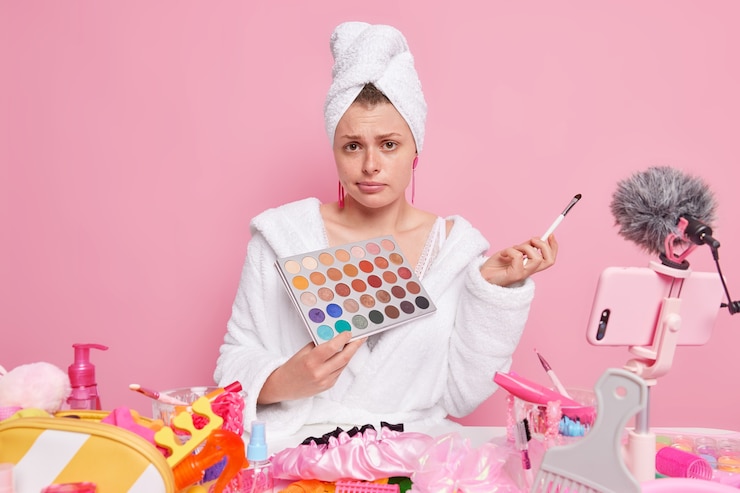Foto gratuito mujer en bata de baño blanca y toalla sostiene una paleta de sombras de ojos y un cepillo cosmético que transmite sobre el maquillaje a los suscriptores aislados en rosa.