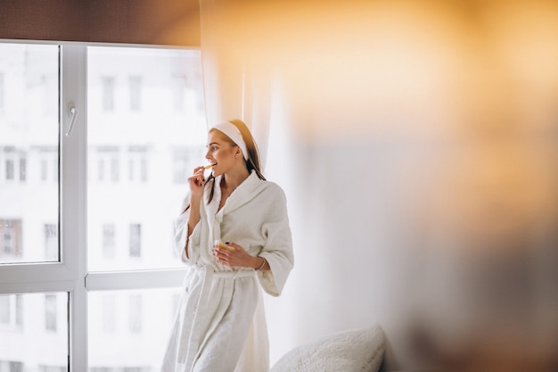 Mujer en bata de baño junto a la ventana y comiendo cereal