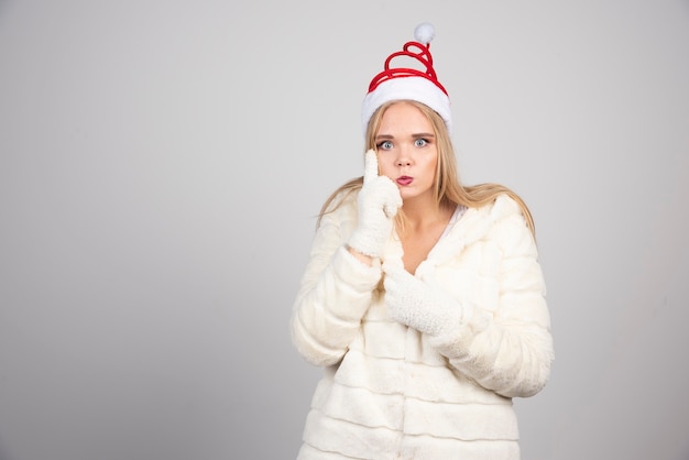 Mujer en bata blanca y gorro de Papá Noel de pie sobre la pared gris.