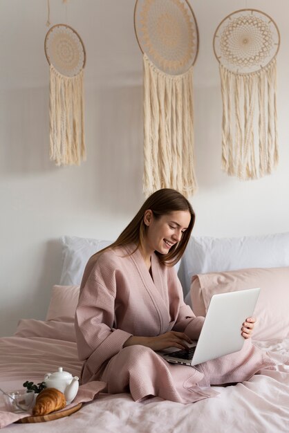 Mujer en bata quedándose en la cama mientras revisa su laptop