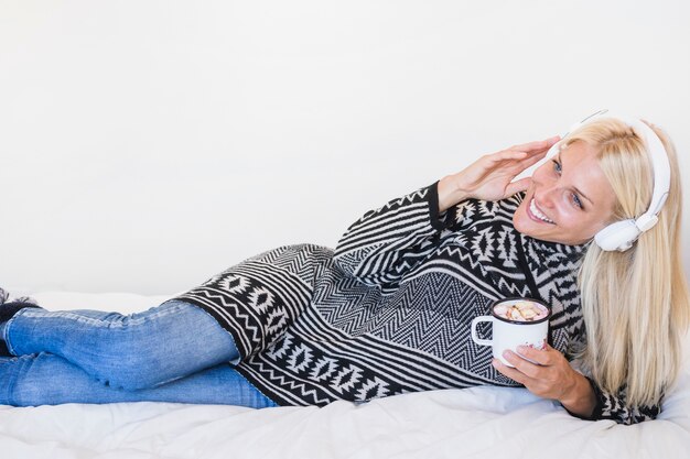 Mujer con bebida caliente disfrutando de la música en la cama