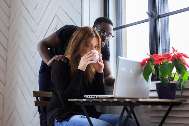 Una mujer bebiendo café por la mañana y un hombre negro anteojos usando una computadora portátil.
