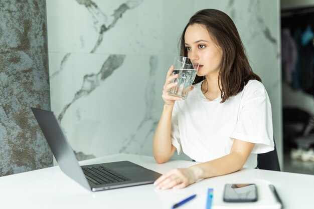 Mujer bebiendo de un vaso de agua mientras escribe en su computadora portátil para mantenerse hidratado mientras trabaja desde casa