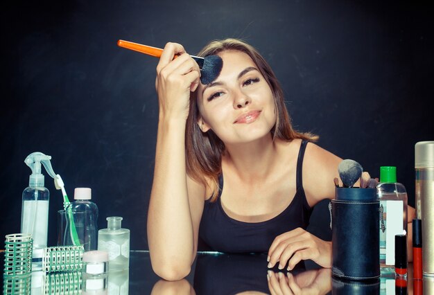 Mujer de belleza aplicando maquillaje. Hermosa chica mirando en el espejo y aplicando cosmético con un pincel grande. Mañana, maquillaje y concepto de emociones humanas. Modelo caucásico en el estudio