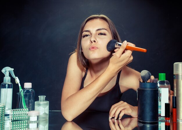 Mujer de belleza aplicando maquillaje. Hermosa chica mirando en el espejo y aplicando cosmético con un pincel grande. Mañana, maquillaje y concepto de emociones humanas. Modelo caucásico en el estudio