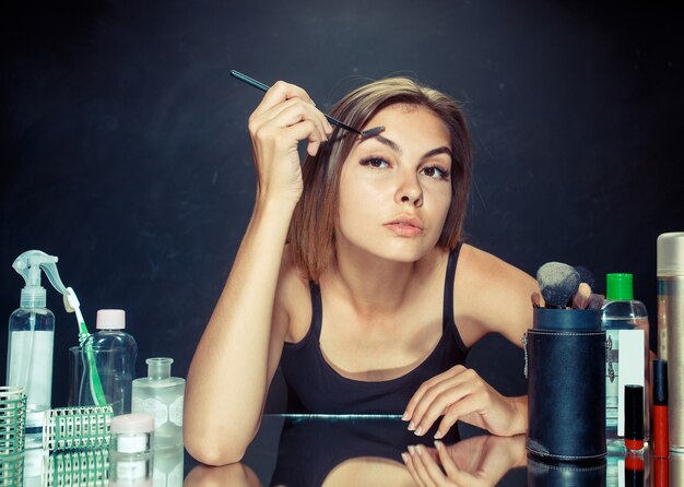 Mujer de belleza aplicando maquillaje. Hermosa chica mirando en el espejo y aplicando cosmético con un pincel grande. Mañana, maquillaje y concepto de emociones humanas.