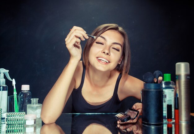 Mujer de belleza aplicando maquillaje. Hermosa chica mirando en el espejo y aplicando cosmético con un pincel