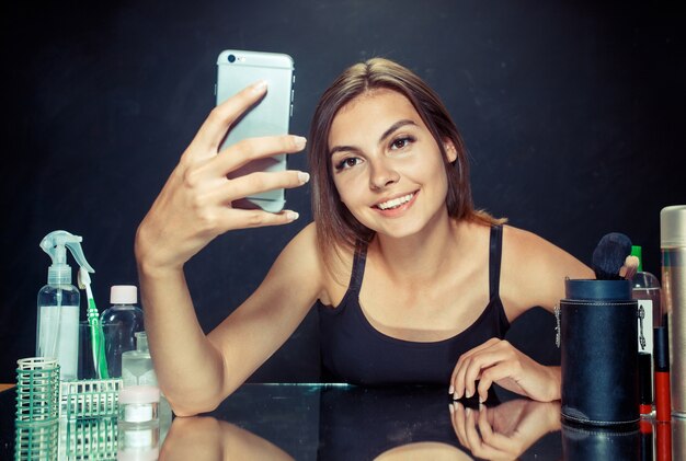 Mujer de belleza después de aplicar el maquillaje. Mujer de belleza con maquillaje. Hermosa chica mirando el teléfono móvil y haciendo foto selfie