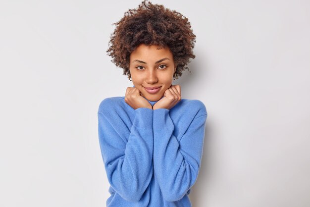 mujer con belleza natural piel sana y radiante mantiene las manos debajo de la barbilla tiene una sonrisa suave en la cara mira directamente a la cámara usa un jersey azul suave posa interior