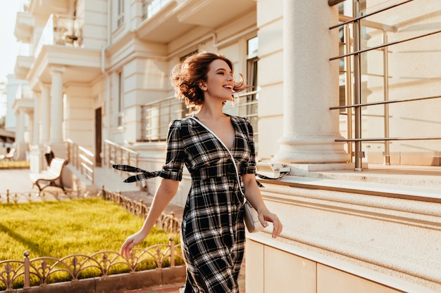 Foto gratuita mujer bien formada en elegante vestido a cuadros caminando por la calle y riendo. preciosa modelo de mujer blanca con corte de pelo corto, pasar tiempo al aire libre.