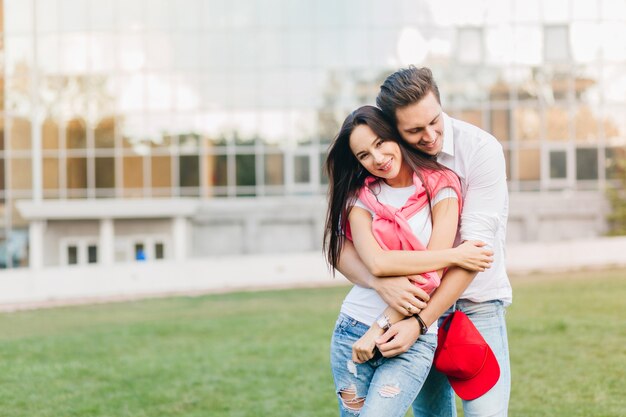 Mujer bien formada en jeans en reloj de pulsera huggs con marido cerca del césped frente al edificio