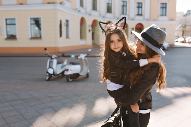 Mujer bien formada de moda con bolsa con niña morena rizada, sonriendo a la cámara. Retrato de joven madre con sombrero elegante pasar tiempo con hermosa hija en fin de semana, ir de compras