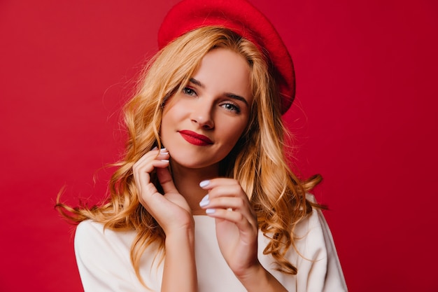 Mujer bien vestida emocional con cabello rubio. Encantadora chica francesa de pie en la pared roja.