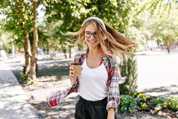 Una mujer blanca alegre bebiendo café al aire libre mientras escucha música Retrato al aire libre de una encantadora chica sonriente con el pelo rubio lacio