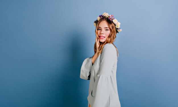 Mujer blanca alegre con hermosas flores en el cabello posando en la pared azul