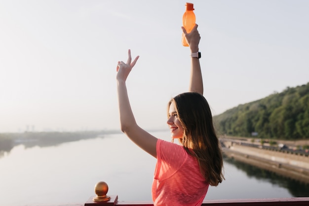 Mujer blanca alegre posando con sonrisa. Retrato al aire libre de chica deportiva inspirada con botella.