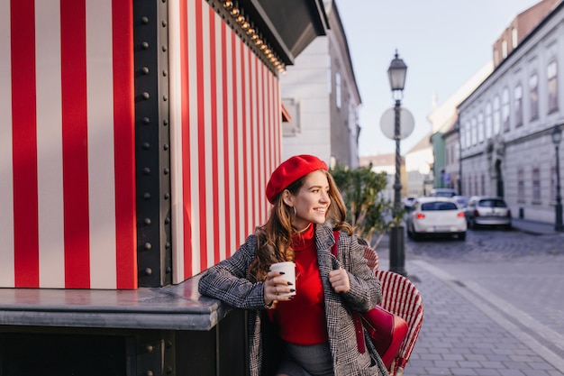 Mujer blanca complacida tomando café en la calle y mirando a su alrededor con linda sonrisa