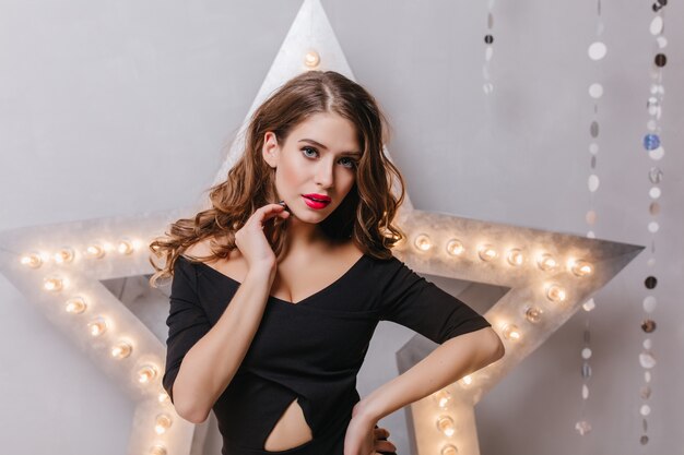 Mujer blanca confiada en traje negro posando junto a la estrella brillante. Foto interior de elegante dama europea con cabello largo castaño mirando con interés.