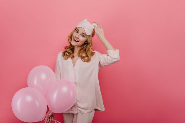 Mujer blanca despreocupada tocando la máscara mientras se ríe en la pared rosa. Bonita cumpleañera en ropa de dormir disfrutando de fiesta con globos.