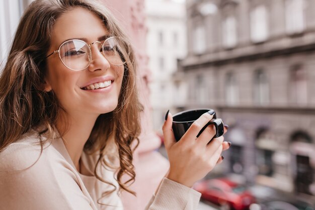 Mujer blanca emocional lleva gafas posando sobre fondo borroso con taza de bebida caliente