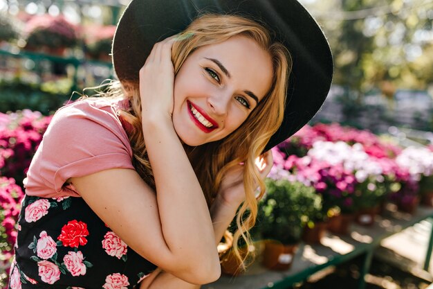 Mujer blanca encantadora que expresa felicidad durante la sesión de retratos con flores. Retrato de encantador modelo caucásico con sombrero sentado en la naturaleza