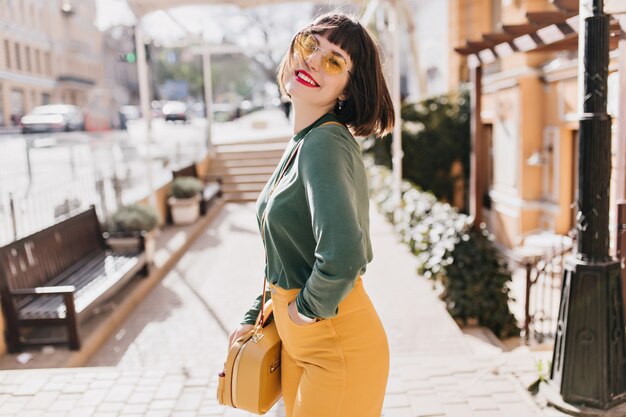 Mujer blanca de ensueño con bolso de moda riendo en la calle urbana. Foto al aire libre de una increíble chica morena con un elegante corte de pelo.