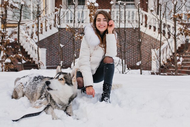 Mujer blanca con increíble sonrisa posando con su perro durante el invierno a pie en el patio. Foto al aire libre de una dama alegre viste pantalones de mezclilla rasgados sentados en la nieve con un husky perezoso.