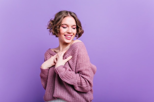 Mujer blanca inspirada viste suéter de punto suave posando con sonrisa tímida. Retrato interior de una joven entusiasta con peinado de moda riendo en la pared púrpura.