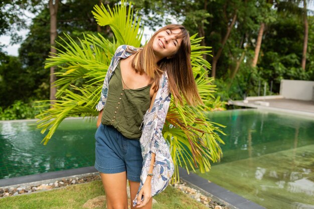 mujer blanca natural posando junto a la piscina y palmeras.