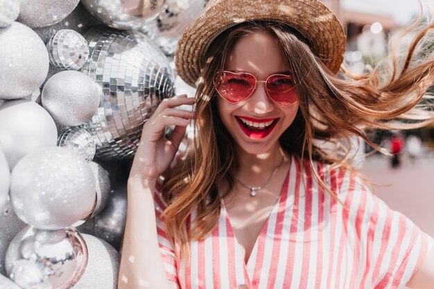 Mujer blanca romántica con largo cabello rubio riendo cerca de bolas brillantes. Adorable niña caucásica con sombrero de paja y gafas de sol rosas disfrutando del verano.