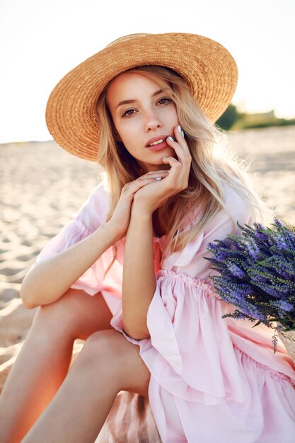 Mujer blanca romántica con sombrero de moda y elegante vestido rosa posando en la playa. Bolsa de paja y ramo de flores.