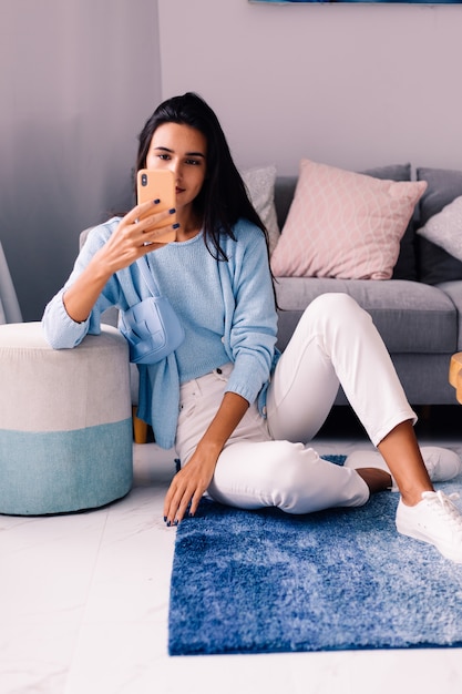 Mujer blogger de moda morena en forma europea se sienta en el piso en la sala de estar cerca del sofá con el teléfono