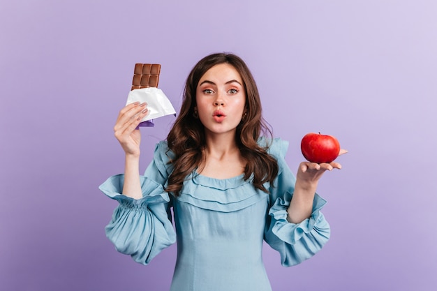 Mujer en blusa azul está posando en la pared púrpura. Chica atractiva piensa en su dieta, eligiendo entre jugosa manzana y chocolate graso.