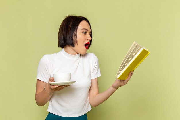 Mujer con blusa blanca y falda verde leyendo un libro con una taza de té