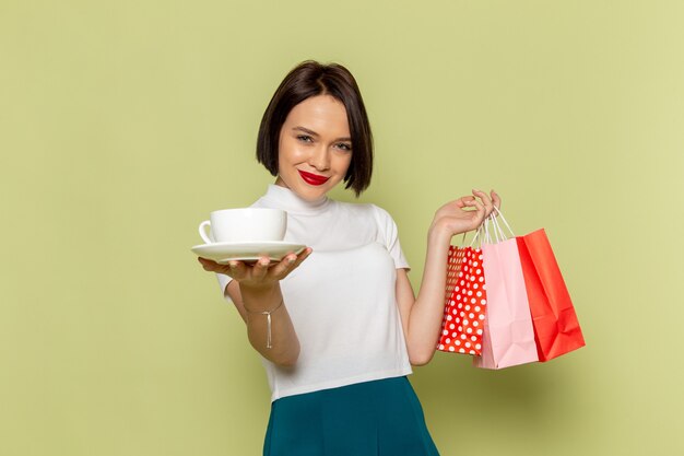 Mujer en blusa blanca y falda verde sosteniendo paquetes de compras y taza blanca