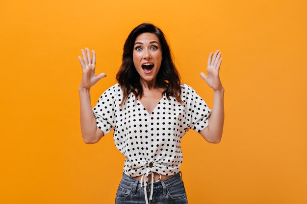 Mujer en blusa ligera grita alegremente y mira a cámara sobre fondo naranja. La chica adulta maravillosa en jeans y camisa de lunares está muy sorprendida.