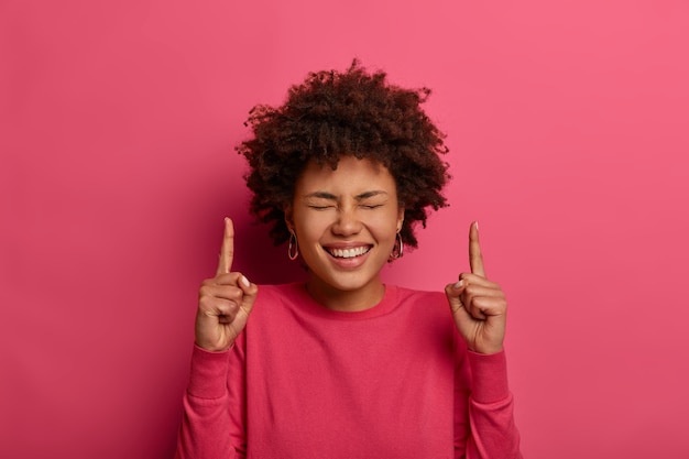 Mujer bonita alegre con expresión de alegría, risas y buen humor, indica los dedos índices hacia arriba, vestida de manera informal, aislada sobre una pared rosa. gente, anuncio, concepto de promoción.