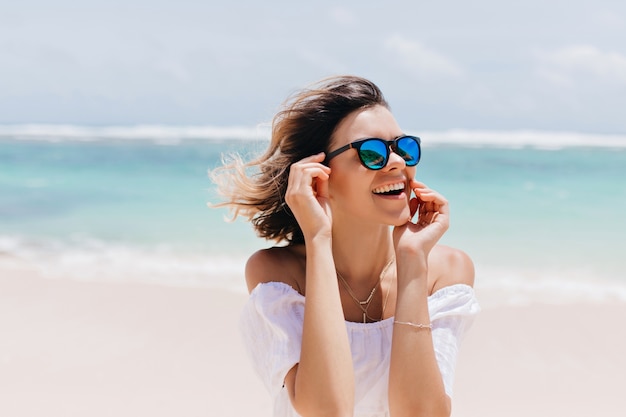 Mujer bonita alegre en gafas de sol brillantes que expresan felicidad en el resort. Tiro al aire libre de la bella dama de buen humor posando en el mar en un día ventoso.