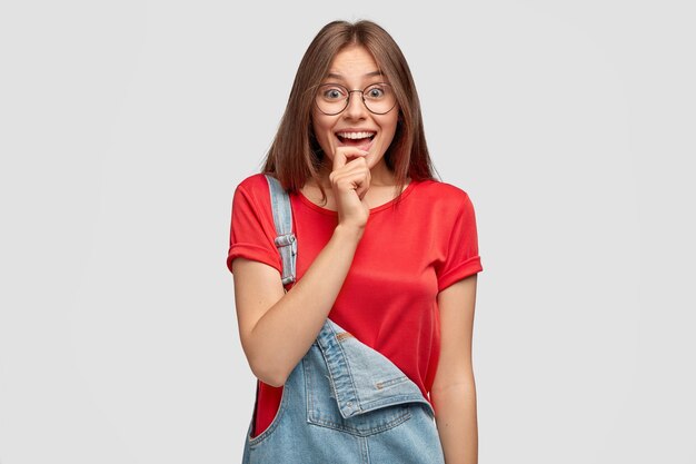 Mujer bonita alegre lleva gafas redondas, mantiene la mano en la mandíbula