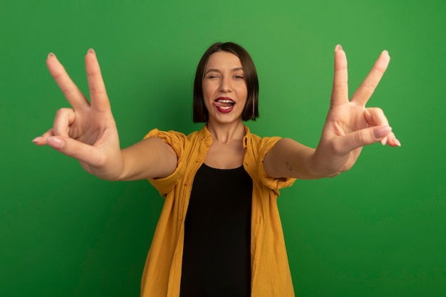 Mujer bonita alegre parpadea el ojo atasca la lengua y hace gestos signo de mano de victoria con las dos manos aisladas en la pared verde