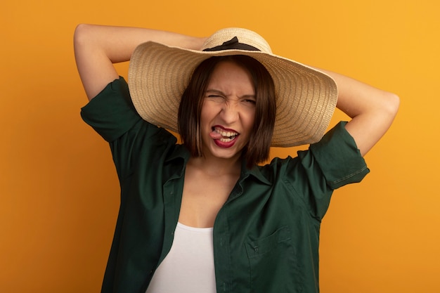 Mujer bonita alegre con sombrero de playa saca la lengua y pone las manos en el sombrero aislado en la pared naranja