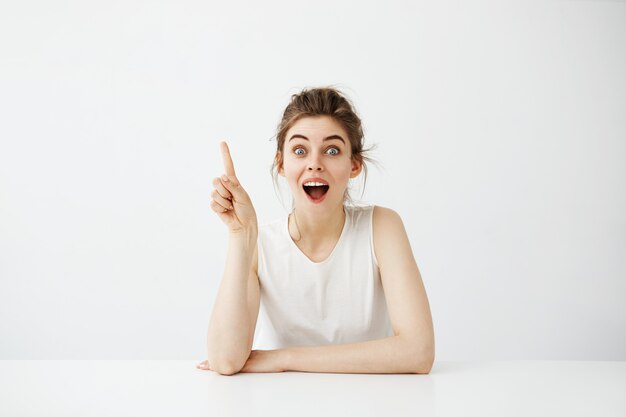 Mujer bonita alegre sorprendida con la boca abierta que señala el dedo que se incorpora en la tabla sobre el fondo blanco.