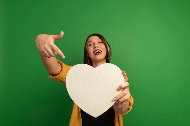 Mujer bonita alegre sostiene y señala en forma de corazón aislado en la pared verde