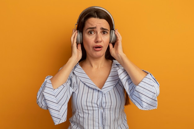 Mujer bonita ansiosa en auriculares mira al frente aislado en la pared naranja