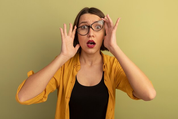 Mujer bonita ansiosa mira al frente a través de gafas ópticas aisladas en la pared verde oliva