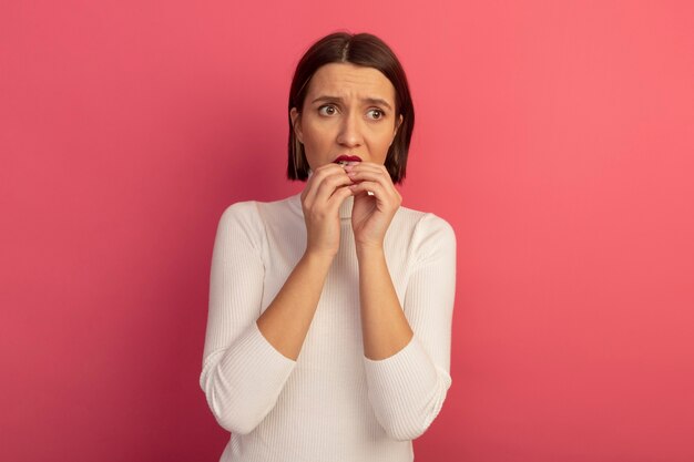 Mujer bonita ansiosa muerde las uñas mirando al lado aislado en la pared rosa